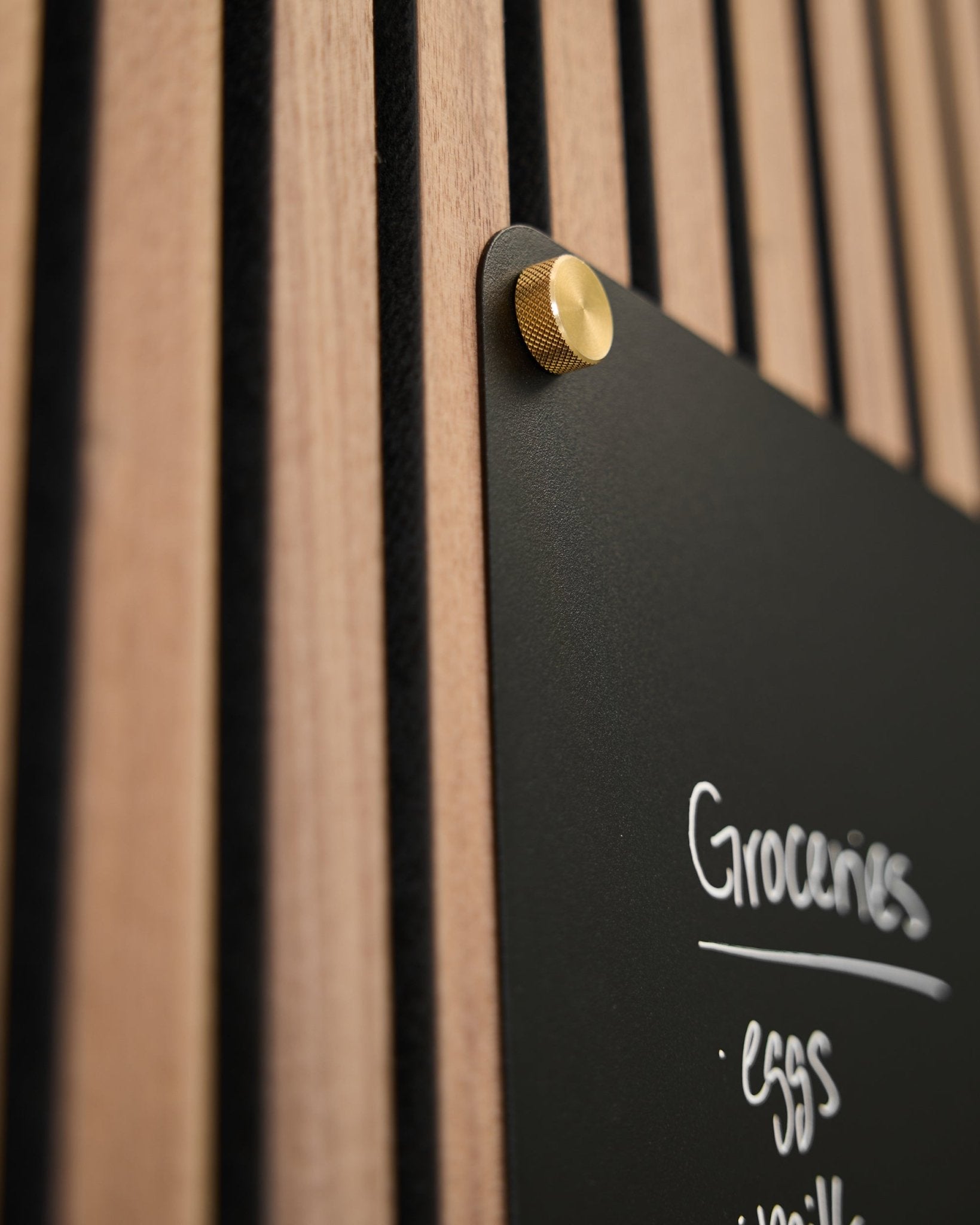 A black board with a handwritten grocery list is mounted to a WoodUpp acoustic slatwall panel using two M5x10 mm brass finger screws.